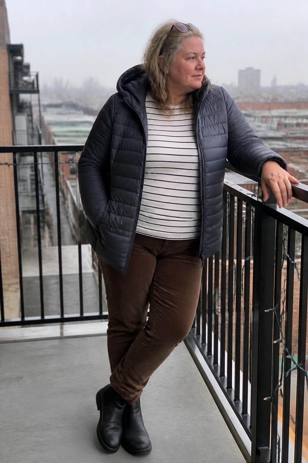 Woman in black puffer jacket, striped shirt, brown corduroy pants on city balcony