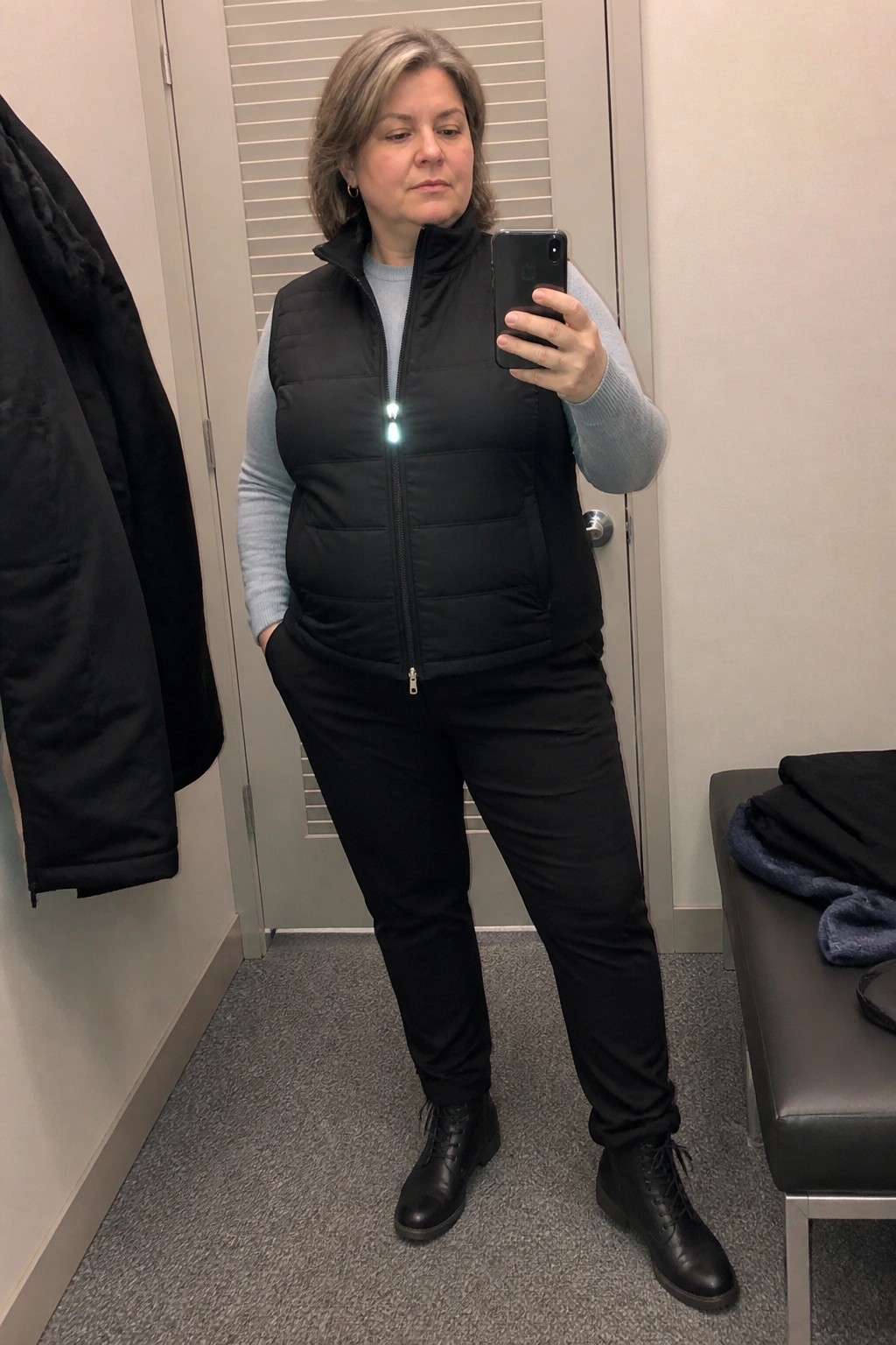 Woman in black puffer vest over blue sweater takes mirror selfie in fitting room.