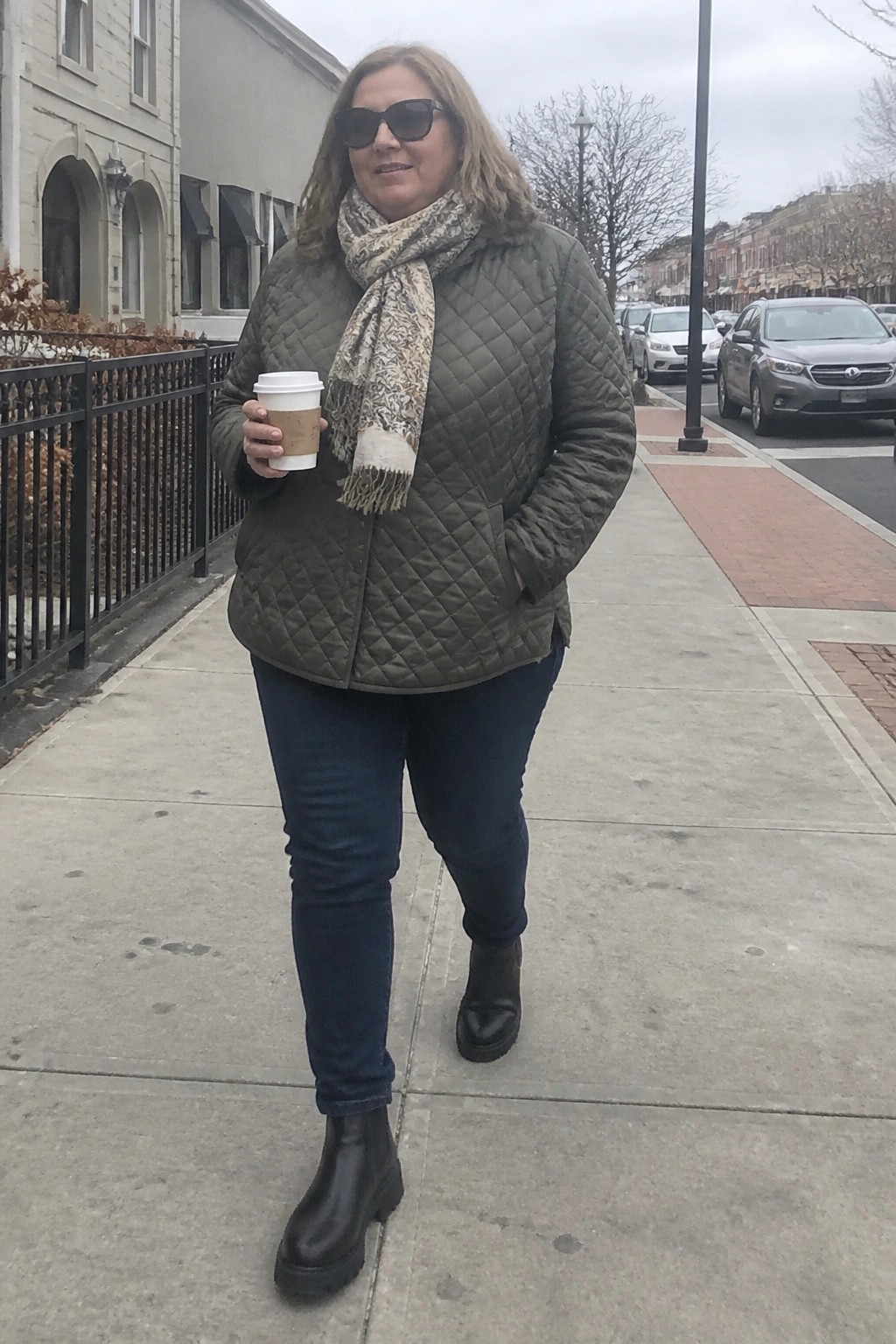 Woman in green quilted jacket and scarf holding coffee on urban sidewalk.