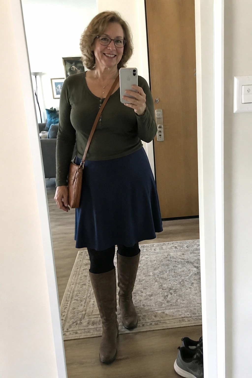 Woman in green top and blue skirt taking mirror selfie with phone and brown bag