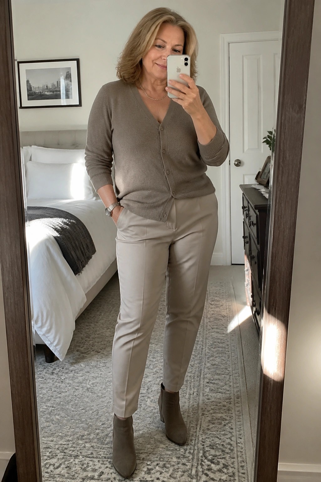Woman in beige cardigan and pants taking mirror selfie in bedroom