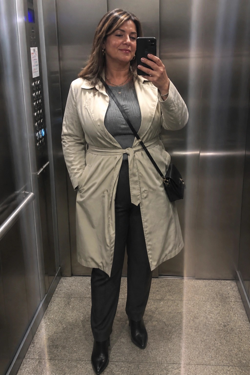 Woman in beige trench coat taking selfie in elevator.