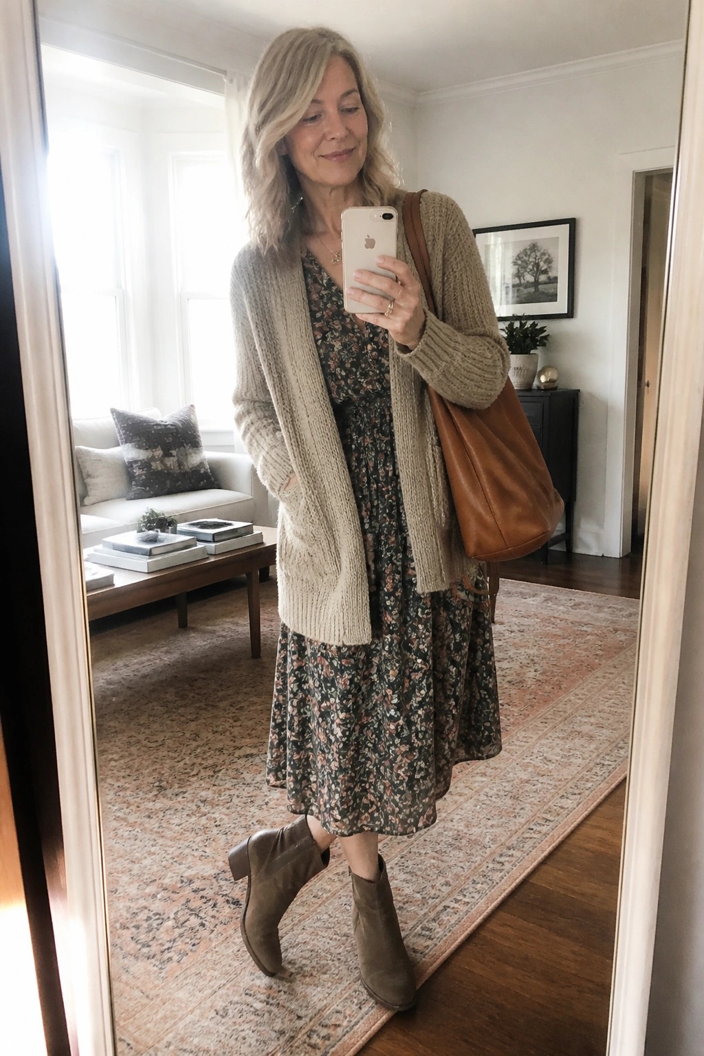 Woman in beige cardigan over floral dress taking mirror selfie with phone
