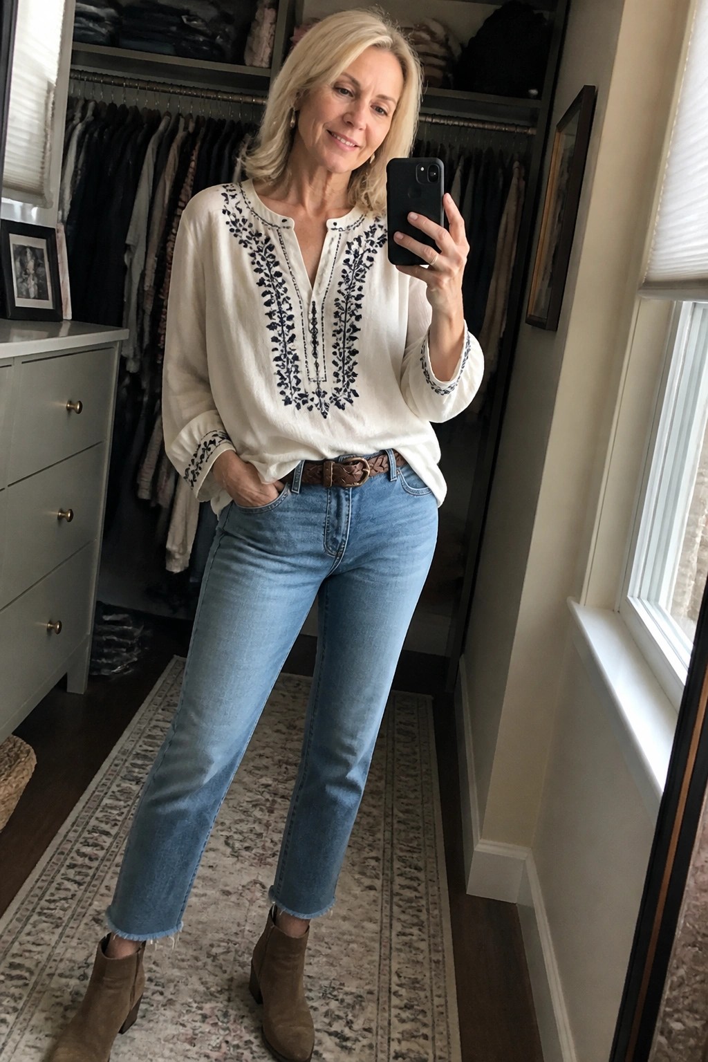 Blonde woman in embroidered white blouse and blue jeans taking mirror selfie in closet