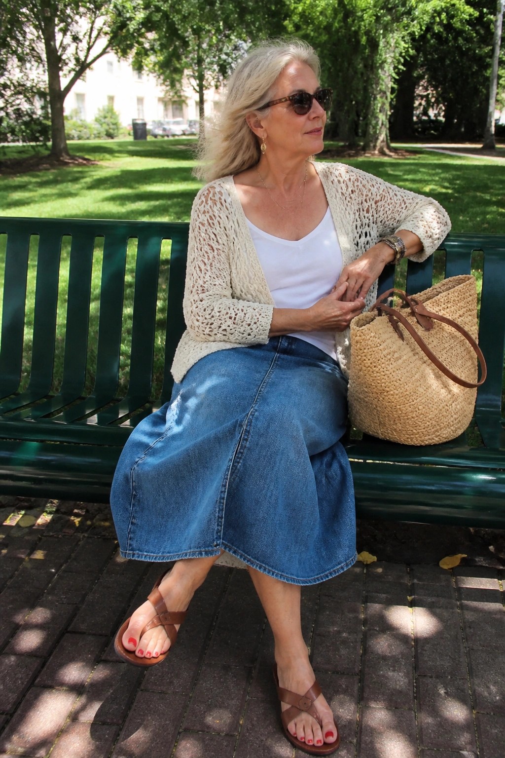Silver-haired woman in denim skirt and knit cardigan sits on green park bench with woven bag.