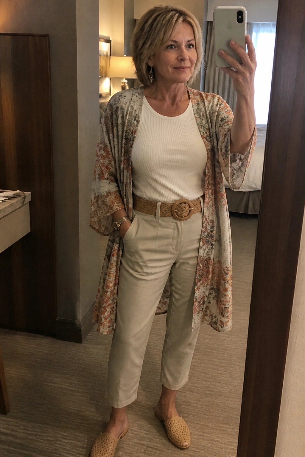 Woman in floral kimono, white top, and beige pants taking mirror selfie in hotel room