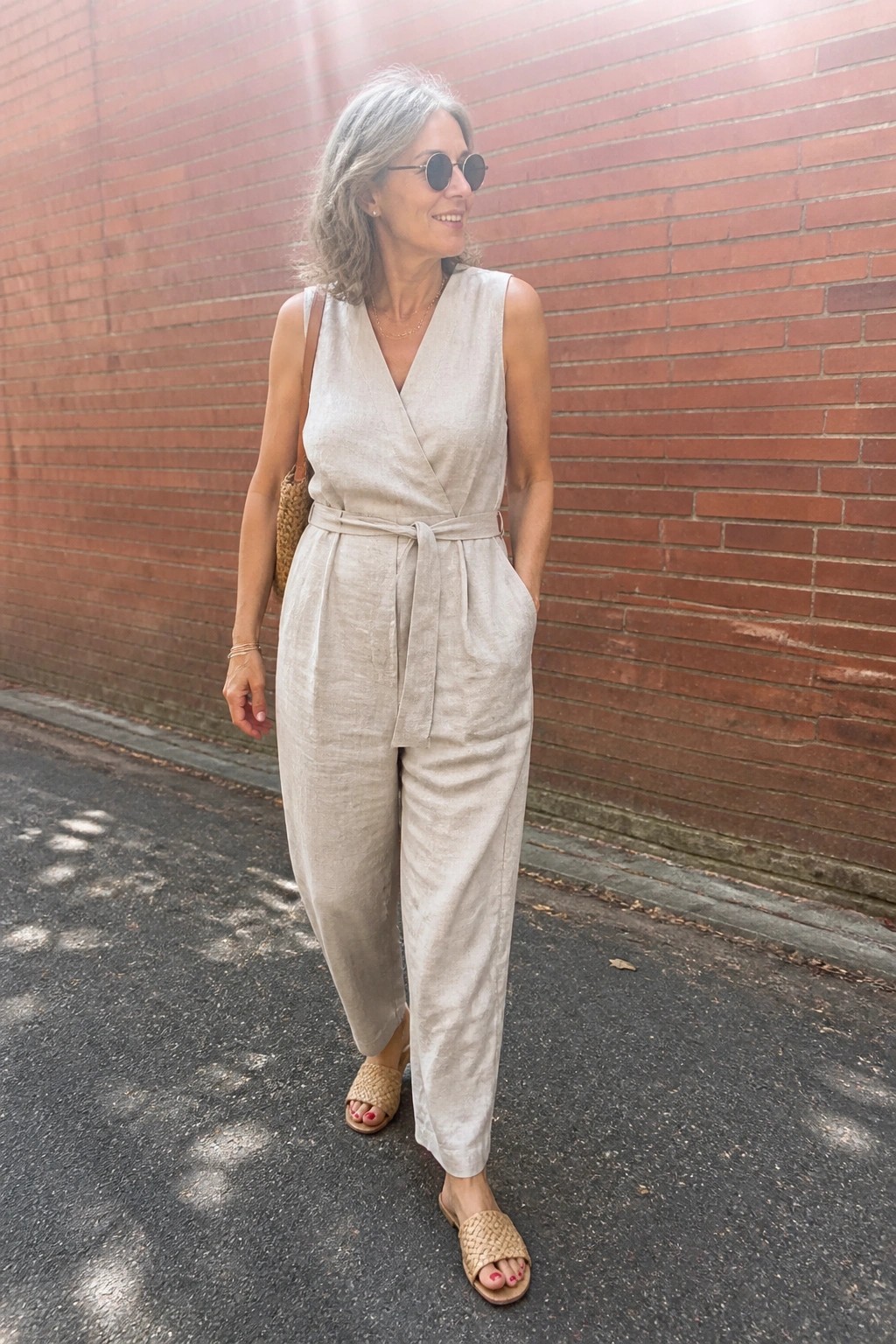 Woman over 50 wears a beige linen sleeveless wrap jumpsuit with tie belt, wide legs, and side pockets, paired with woven slide sandals, a woven shoulder bag, round sunglasses, and layered necklaces.