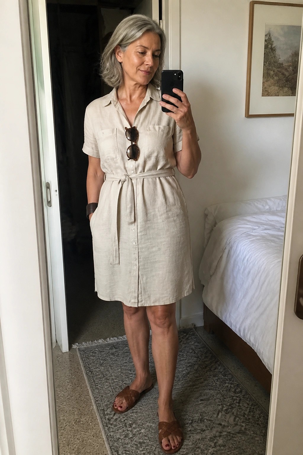 Woman over 50 in a light beige linen shirt dress with short sleeves tie belt and front pockets paired with brown woven slides for a mirror selfie.