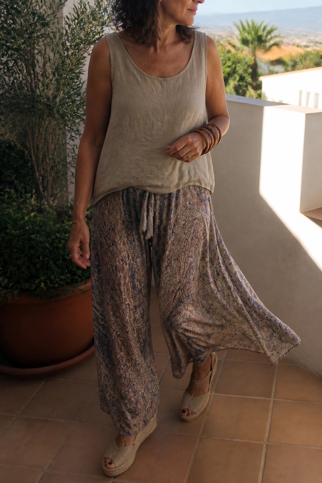 Woman over 50 wearing a beige linen tank top tucked loosely into wide leg patterned pants with a tie waist, paired with beige wedge espadrille sandals and stacked brown bracelets on one wrist.