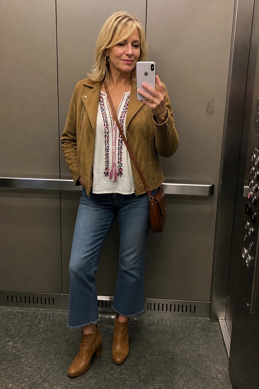 Blonde woman in brown suede jacket and embroidered top taking elevator selfie