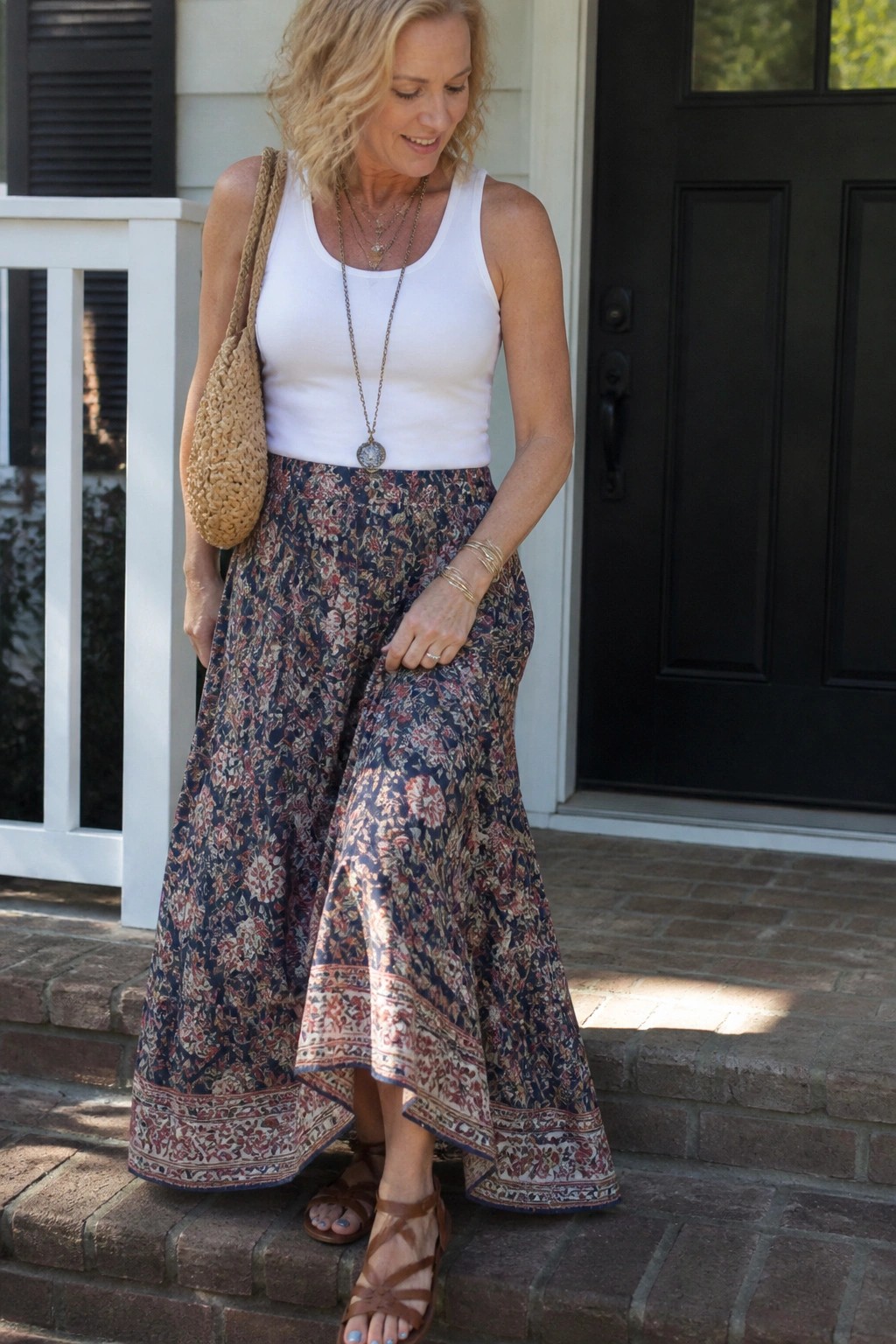 Blonde woman in white tank top and floral maxi skirt holding woven bag on steps.