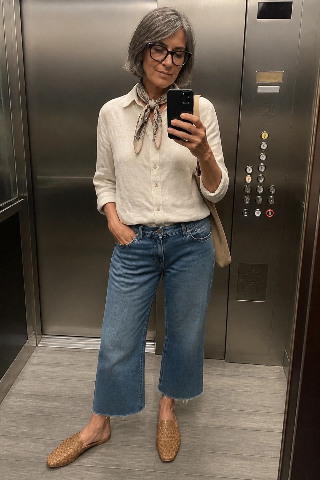 Woman over 50 wearing a white button down shirt tied with a patterned scarf, blue wide leg cropped jeans, and woven brown mules while taking a mirror selfie in an elevator.