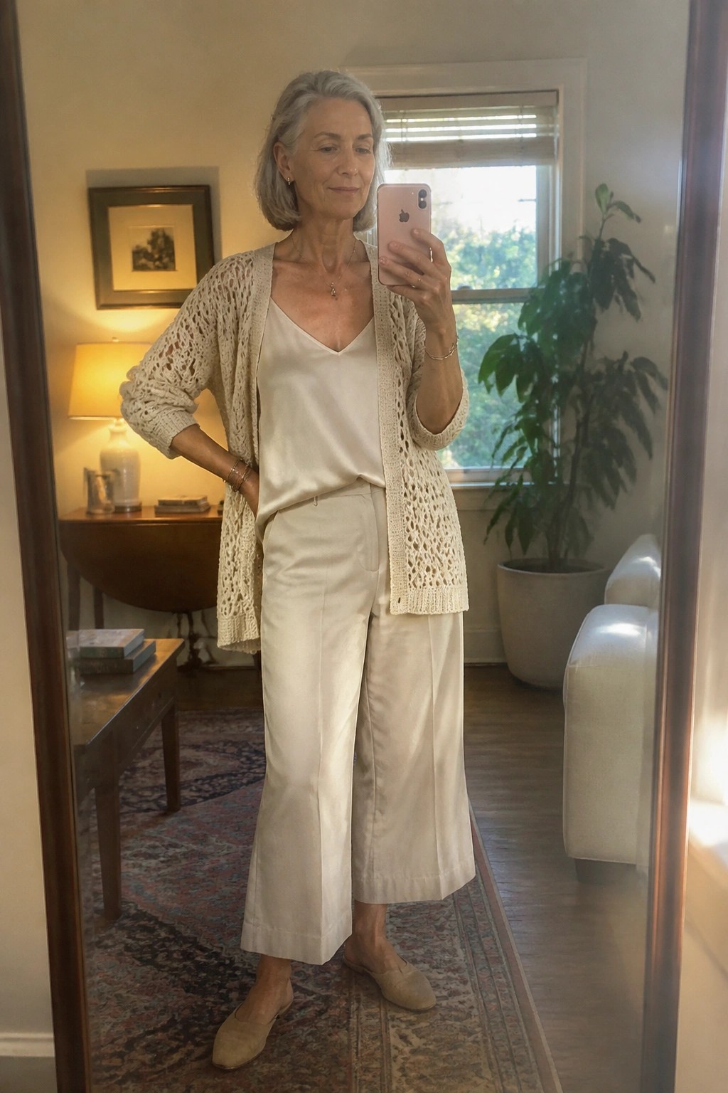 Gray-haired woman in cream cardigan and wide pants taking mirror selfie with phone