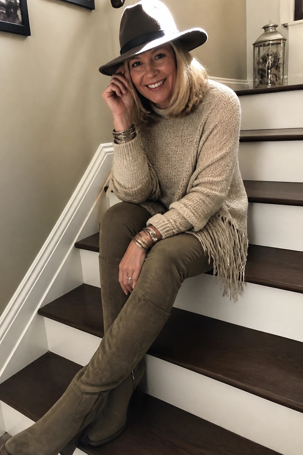 Blonde woman in fringed beige sweater and olive boots seated on wooden stairs wearing wide hat