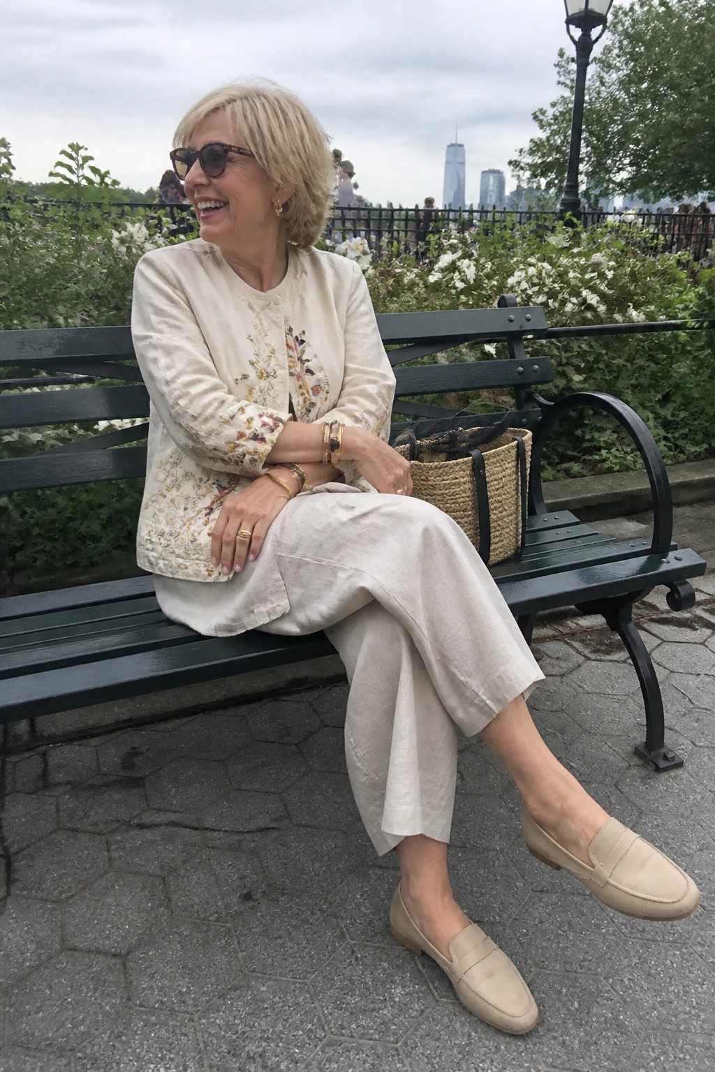 Blonde woman in cream floral jacket and pants smiles on park bench with straw bag.