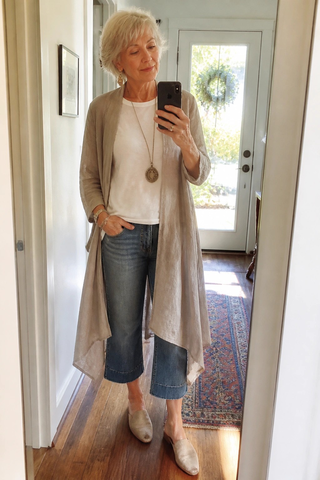 White-haired woman in beige cardigan and cropped jeans taking mirror selfie in hallway.