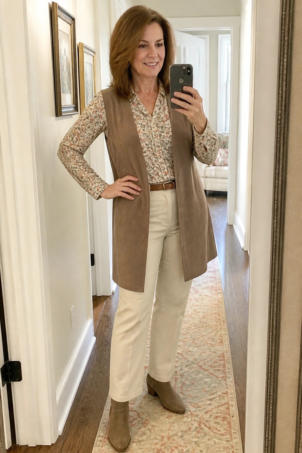Woman in floral blouse, brown suede vest, and white pants taking mirror selfie