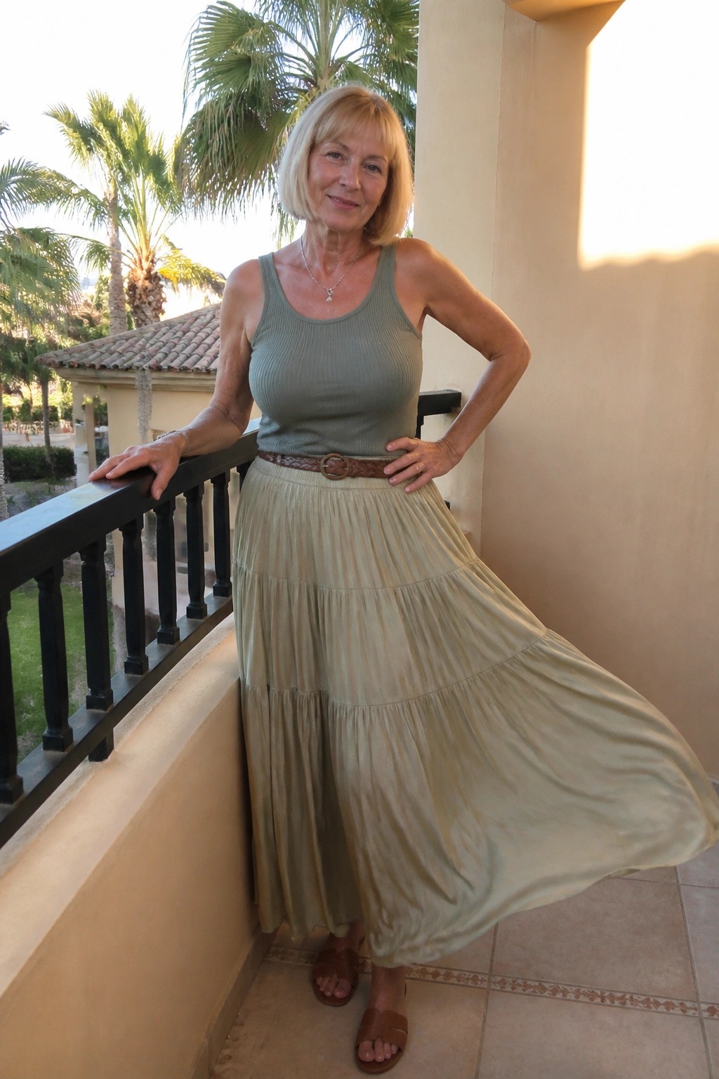 Blonde woman in sage tank top and tiered beige skirt on sunny palm balcony