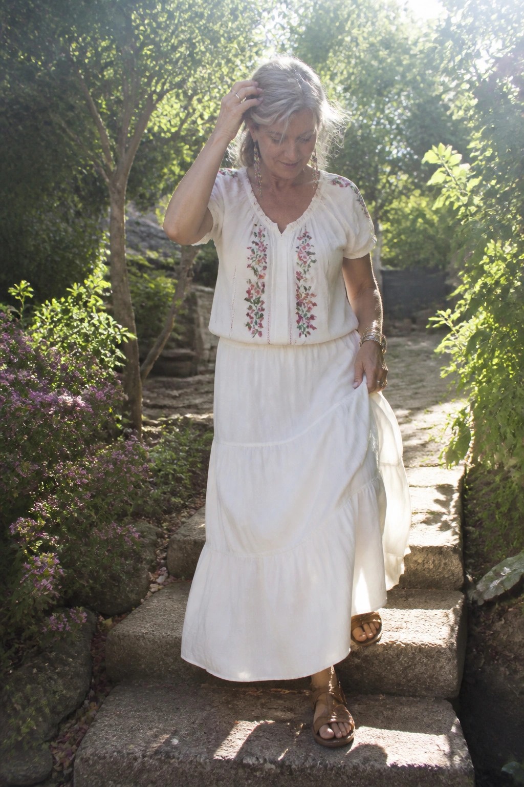 Woman over 50 in white floral embroidered peasant blouse tucked into a tiered white maxi skirt with brown leather sandals and layered bracelets