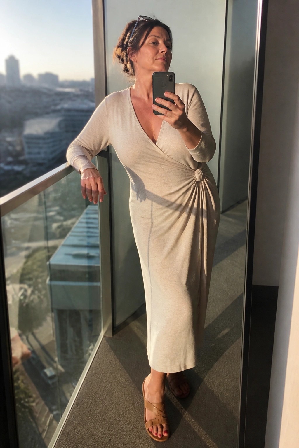 Woman in beige wrap dress taking selfie on sunny city balcony.