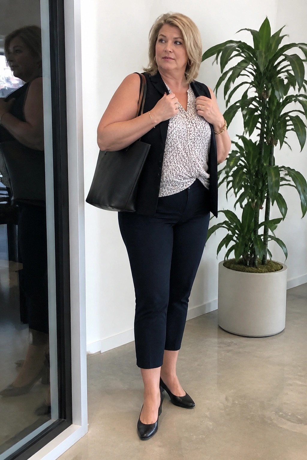 Blonde woman in black vest and pants adjusting outfit beside potted plant.