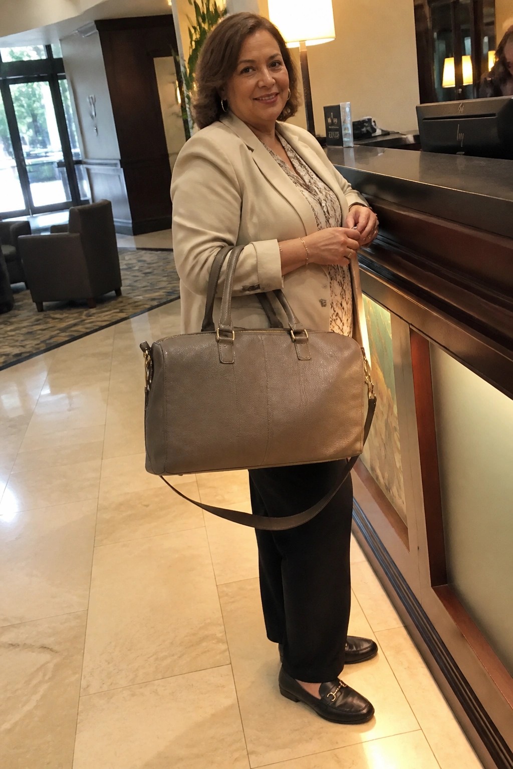 Woman in beige blazer holds gray handbag at hotel reception counter