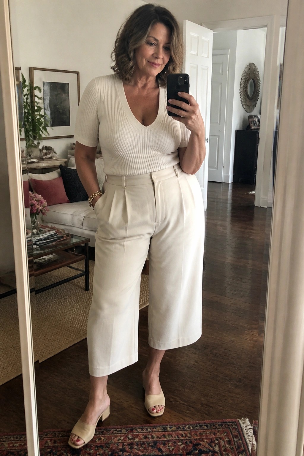 Woman in cream ribbed sweater and white wide-leg pants taking mirror selfie indoors.