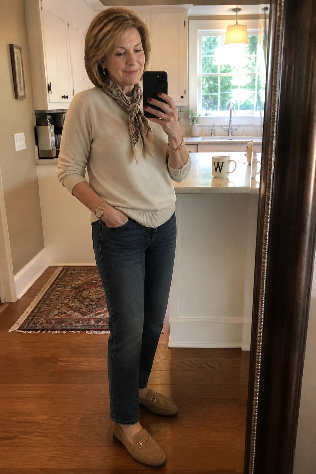 Woman over 50 wearing a cream knit sweater tied with a patterned scarf blue jeans and beige suede loafers standing in a kitchen holding a phone for a mirror selfie.