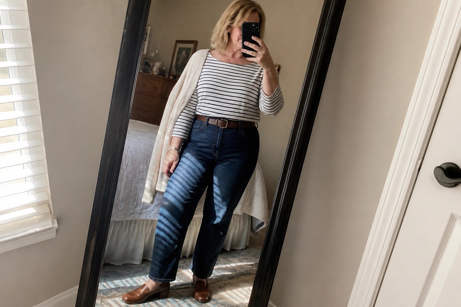 Woman taking mirror selfie in black and white striped long sleeve top layered with beige vest dark blue jeans brown belt and black loafers