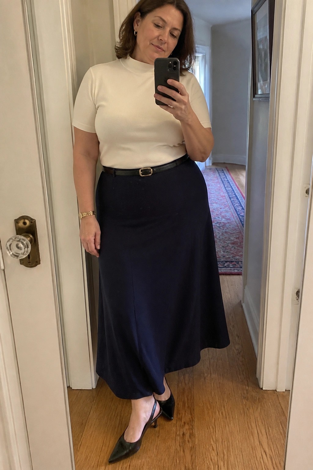 Woman wearing a cream short sleeve top tucked into a navy midi skirt with a thin black belt, black slingback heels, and a gold bracelet while holding a phone for a mirror selfie.