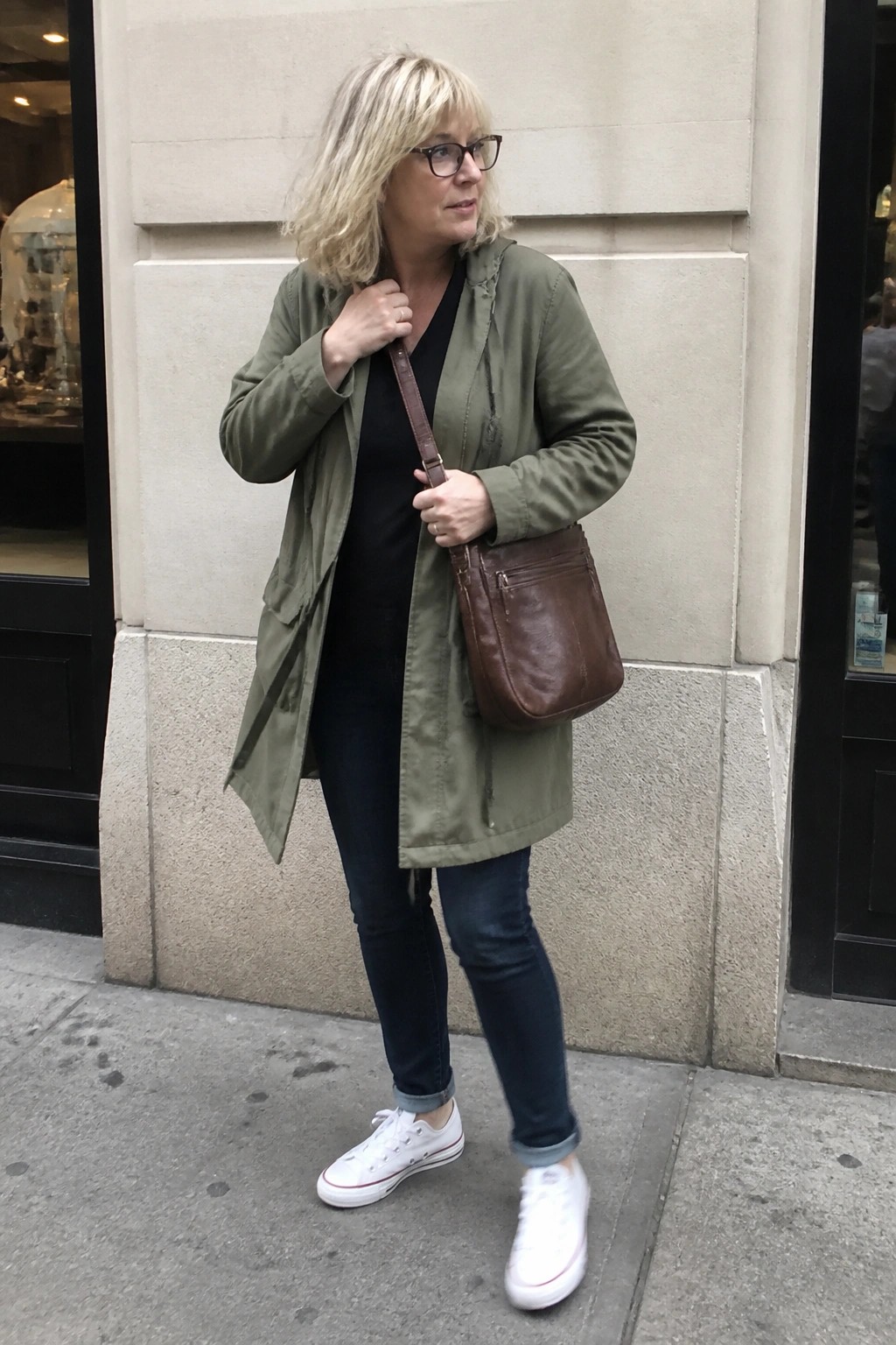 Blonde woman in olive jacket, jeans, and white sneakers with brown bag on sidewalk