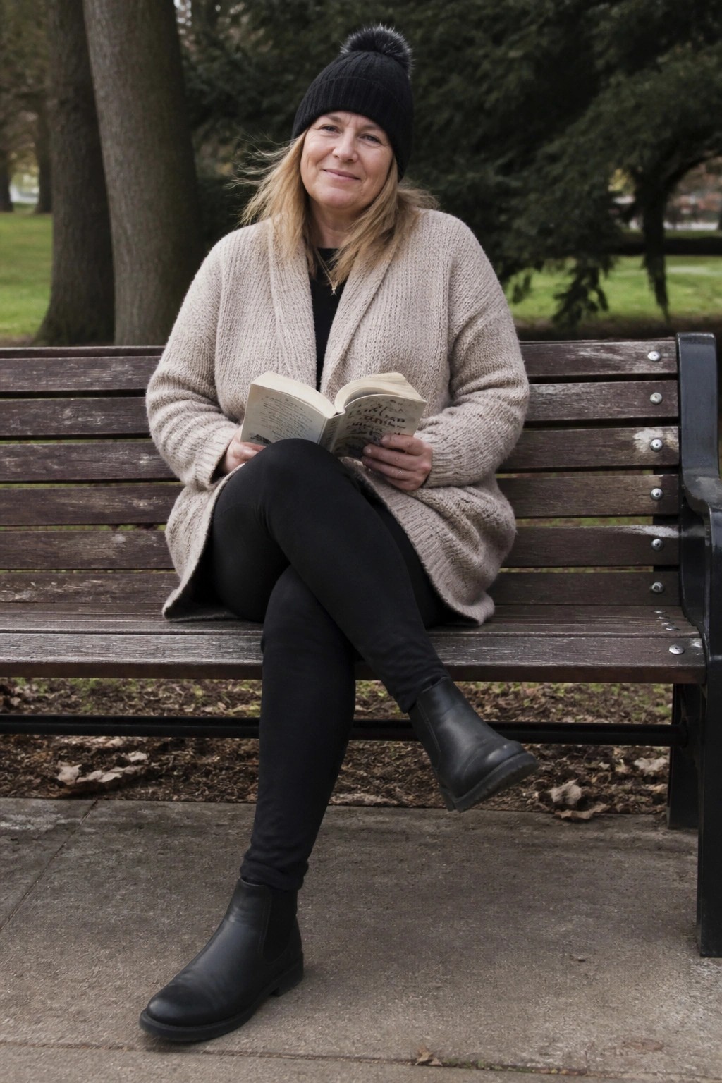 Woman in black beanie and beige cardigan reads book on park bench