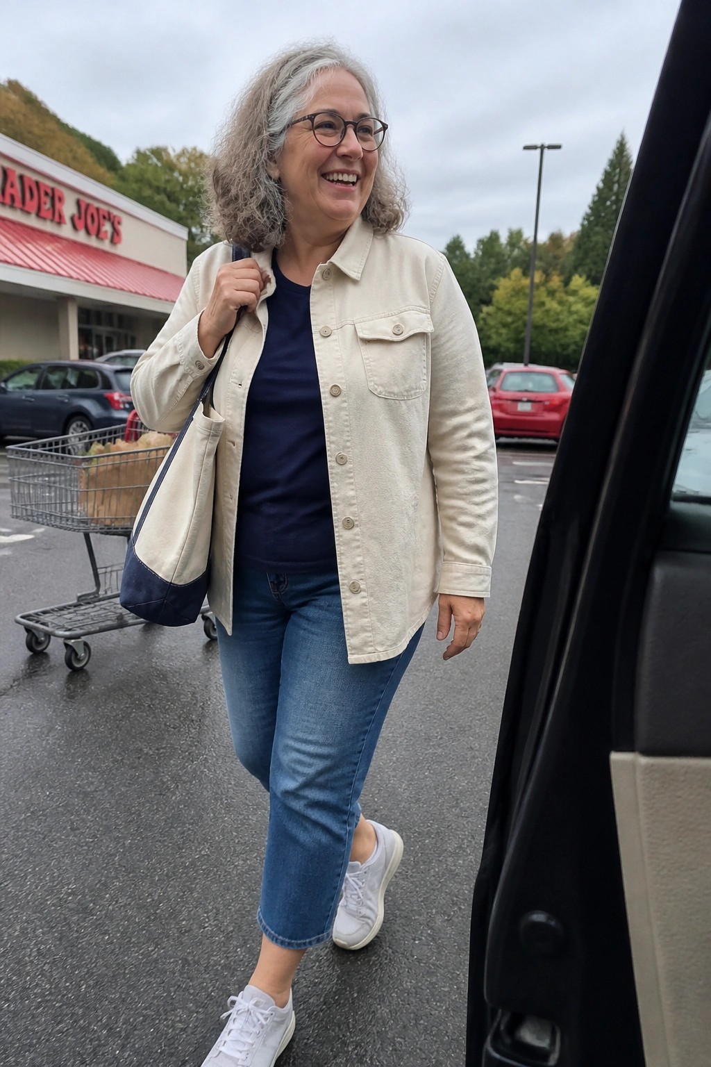 Smiling gray-haired woman in beige jacket and jeans by open car door in parking lot