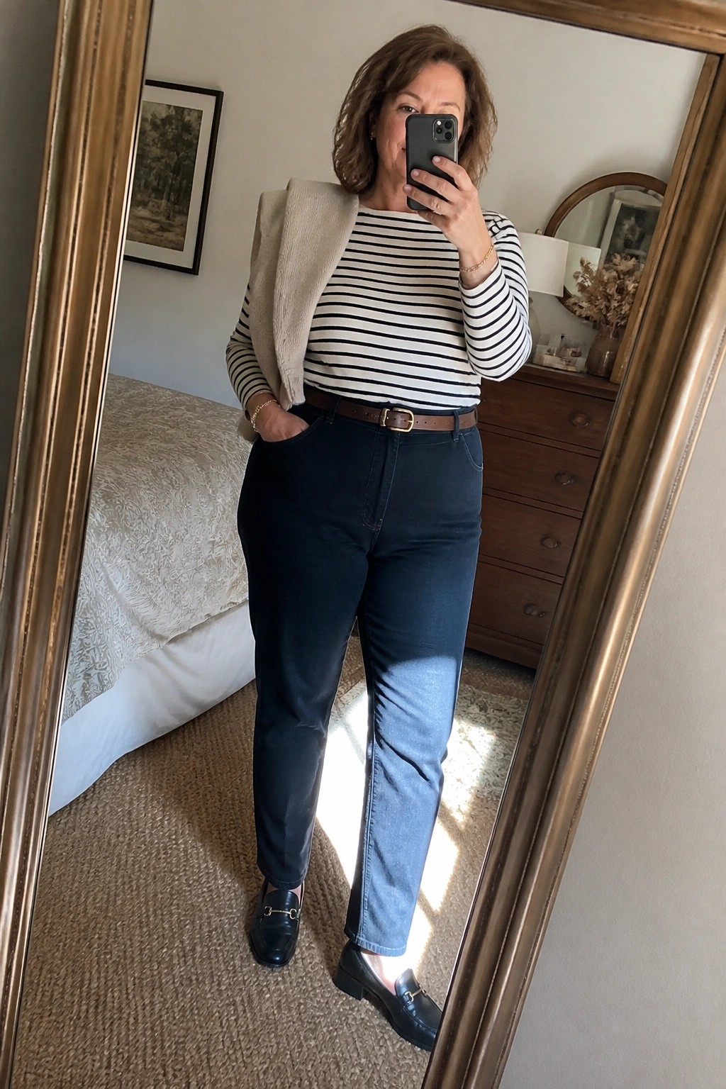 Woman taking mirror selfie in black and white striped long sleeve top layered with beige vest dark blue jeans brown belt and black loafers