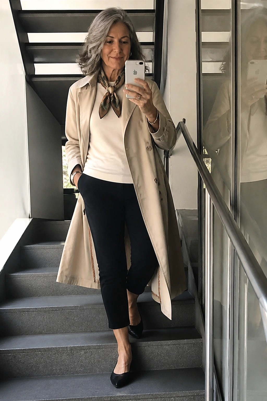 Gray-haired woman in beige trench coat holds white phone on gray staircase.