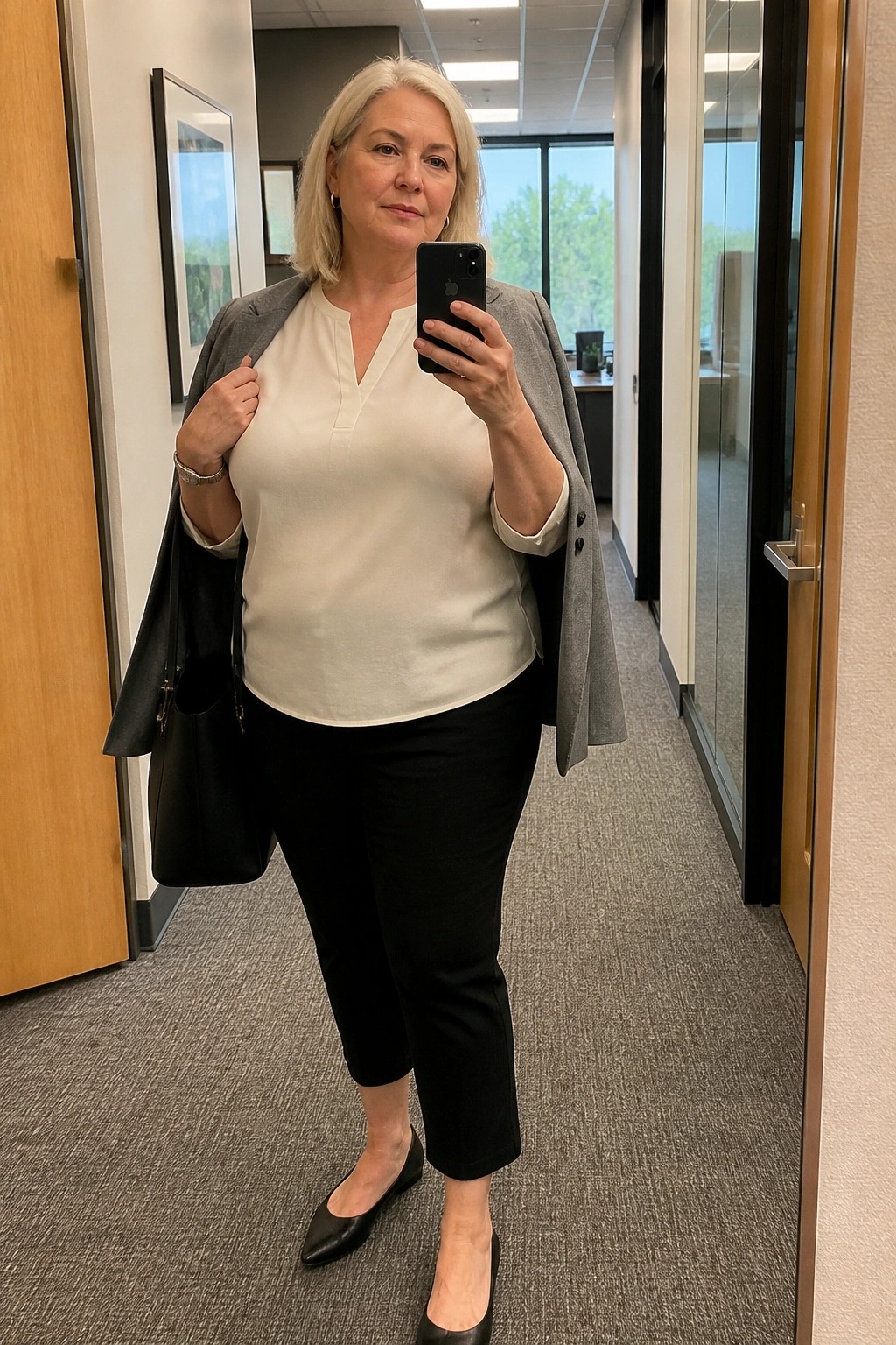 Blonde woman in white blouse and black pants holding phone and gray blazer in office hallway.