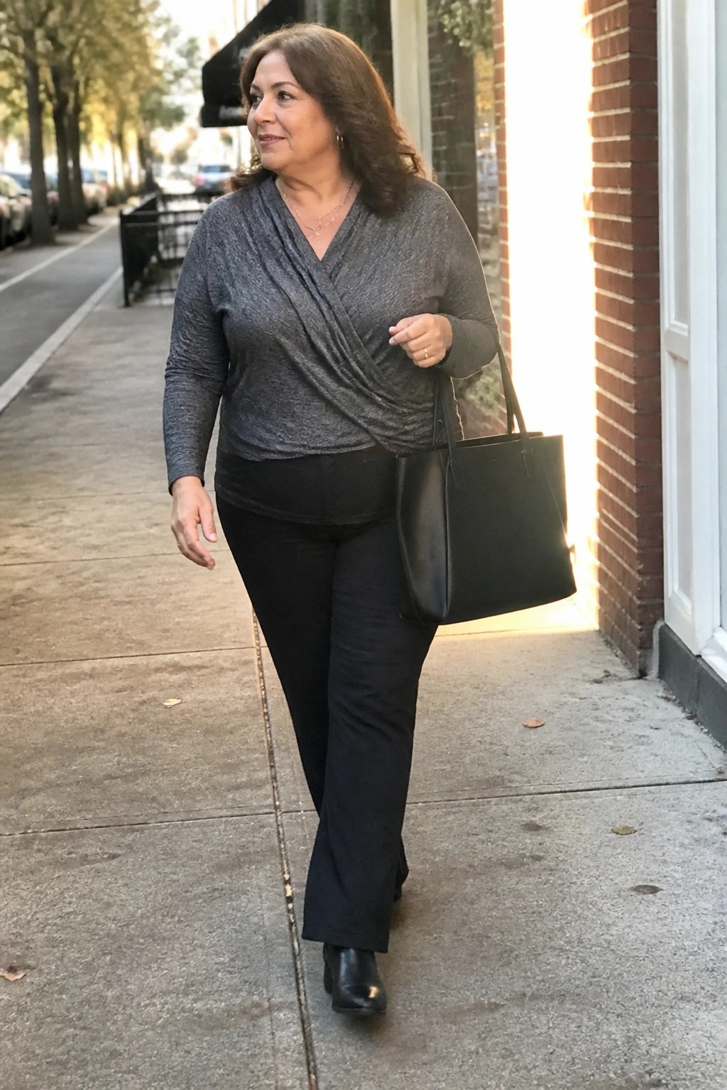 Woman wearing black pull-on jeans with a gray marled wrap top, black ankle boots, and carrying a black leather tote bag on a city sidewalk.