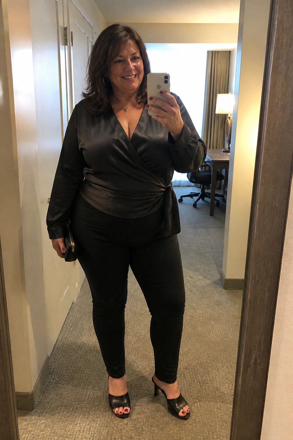 Woman in black satin wrap blouse and pants taking mirror selfie with iPhone