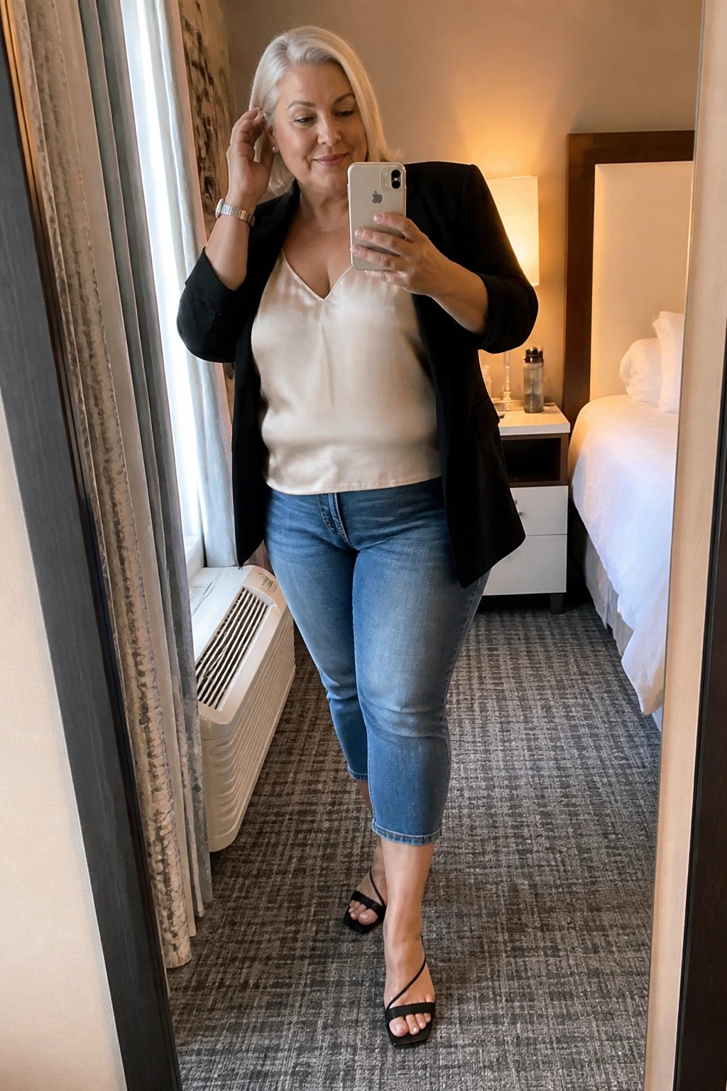 White-haired woman in black blazer and blue jeans taking hotel mirror selfie