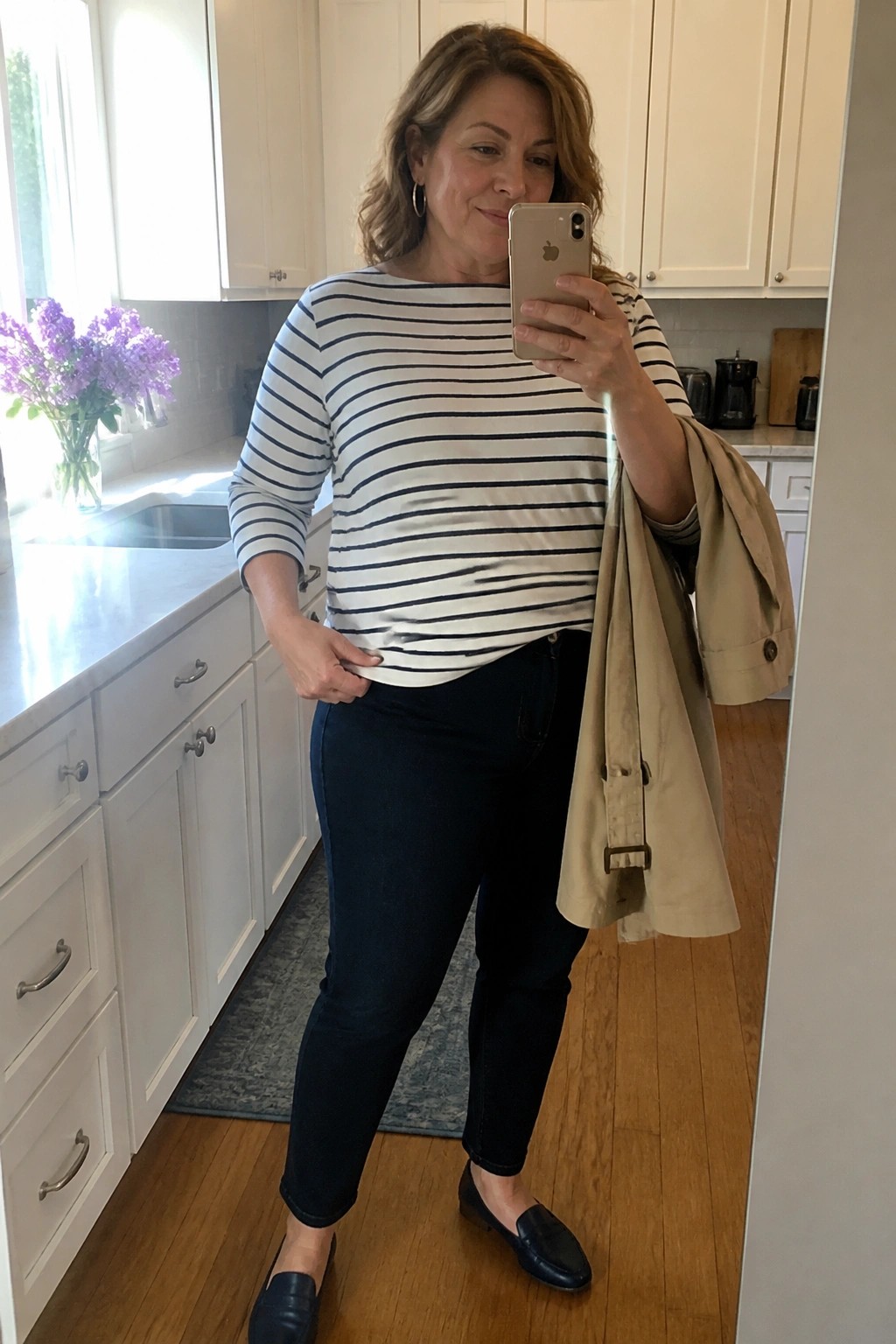 Woman in striped top and jeans taking mirror selfie while holding beige coat