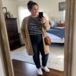 Blonde woman in striped shirt and tan cardigan taking bedroom mirror selfie with phone.