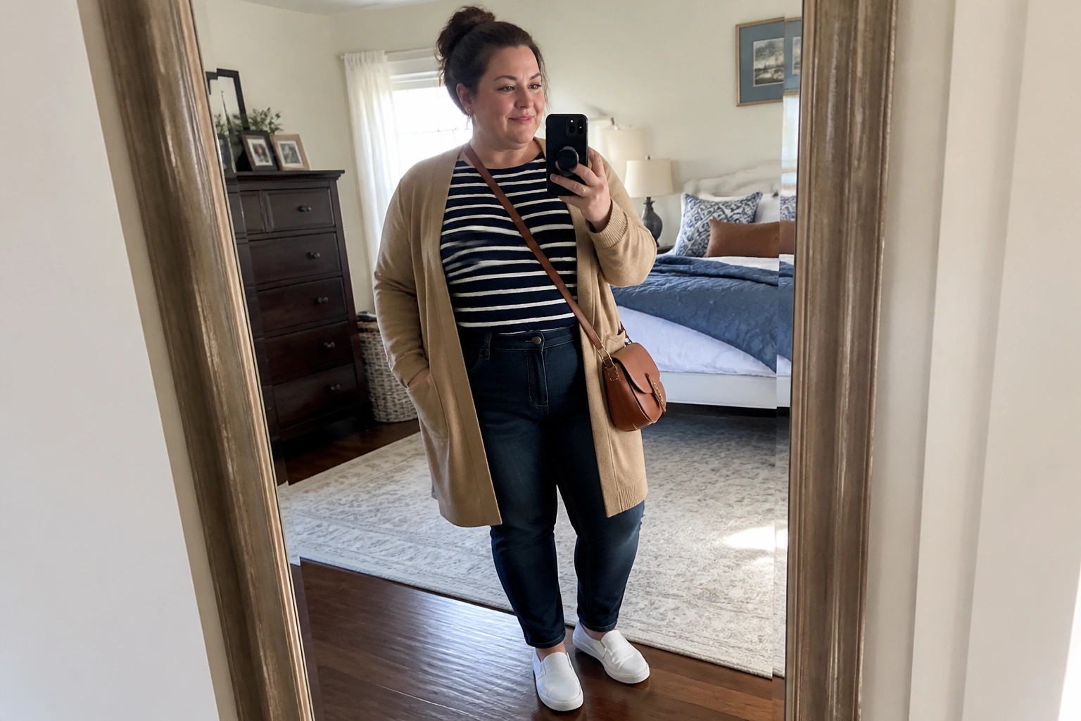 Blonde woman in striped shirt and tan cardigan taking bedroom mirror selfie with phone.