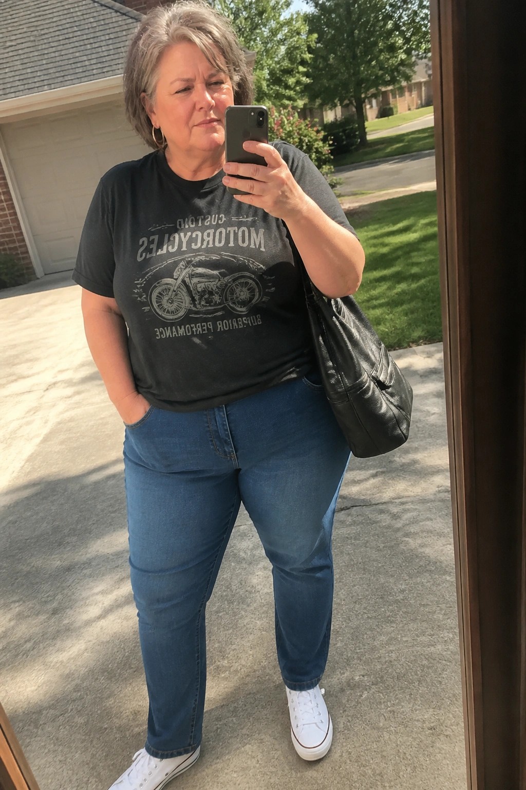 Gray-haired woman in black motorcycle t-shirt, blue jeans, white sneakers taking selfie outdoors
