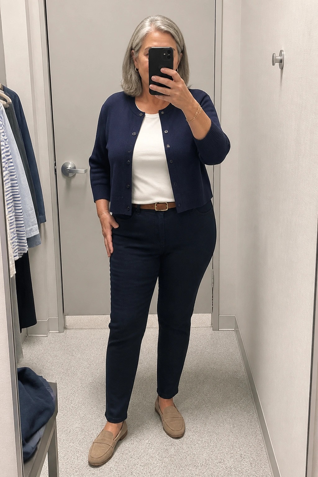 Gray-haired woman in navy cardigan, white top, dark pants taking mirror selfie.