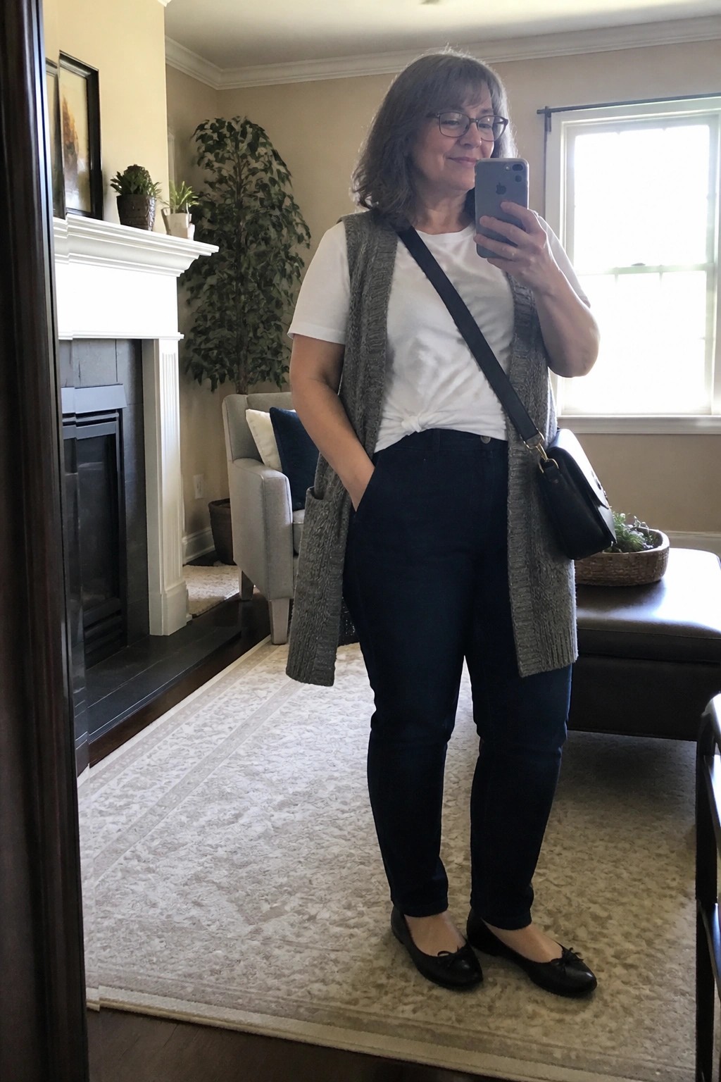 Woman in gray knit vest, white tee, and dark jeans taking mirror selfie in living room