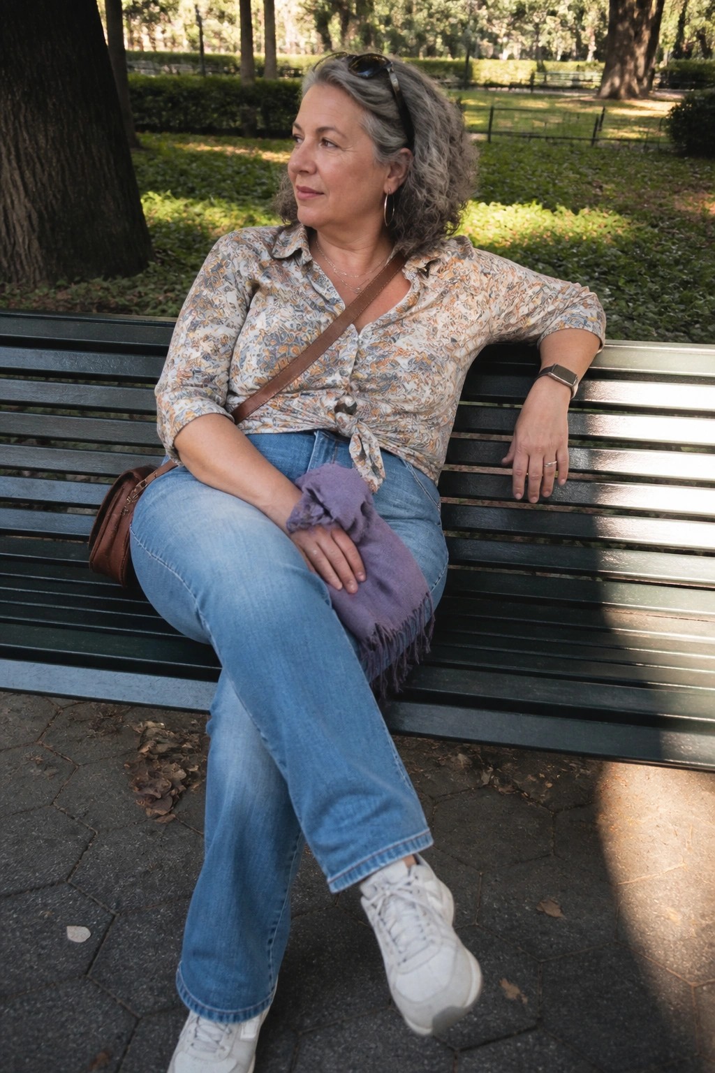 Woman with gray curls in patterned shirt and jeans sits on green park bench with purple shawl