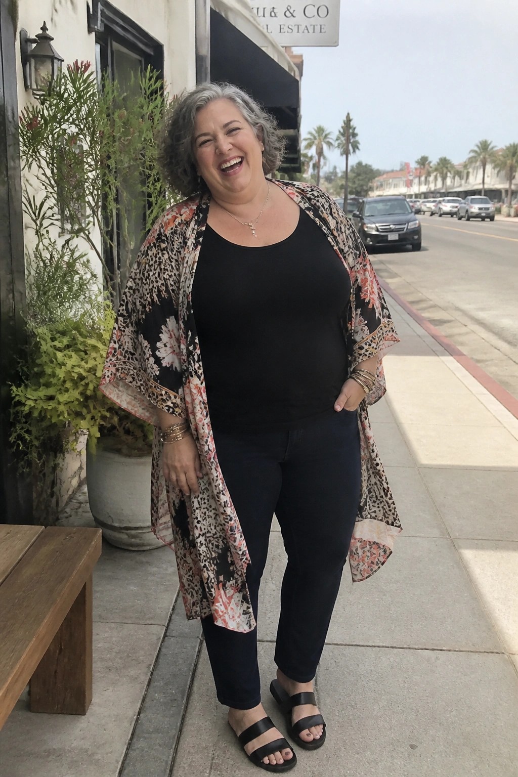 Plus size woman with gray curly hair wearing black pull on jeans, black tank top, open patterned kimono duster, black slide sandals and layered gold jewelry standing outdoors.