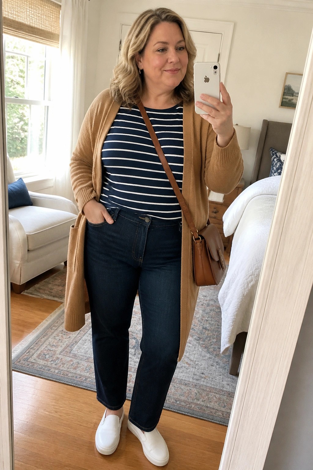 Blonde woman in striped shirt and tan cardigan taking bedroom mirror selfie with phone.